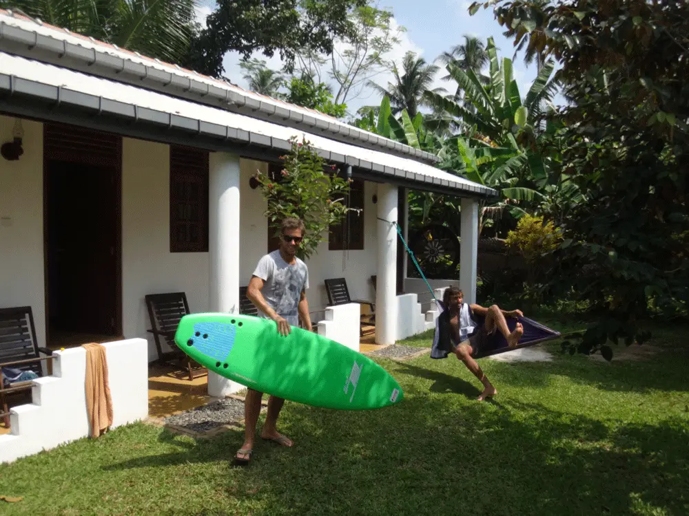 A guest carrying a green soft-top surfboard across the garden in front of the rooms at Surfing Life